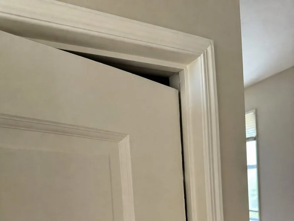 An interior door hanging out of square in its frame, with a tapering wedge of gap open along the head and the latch side.