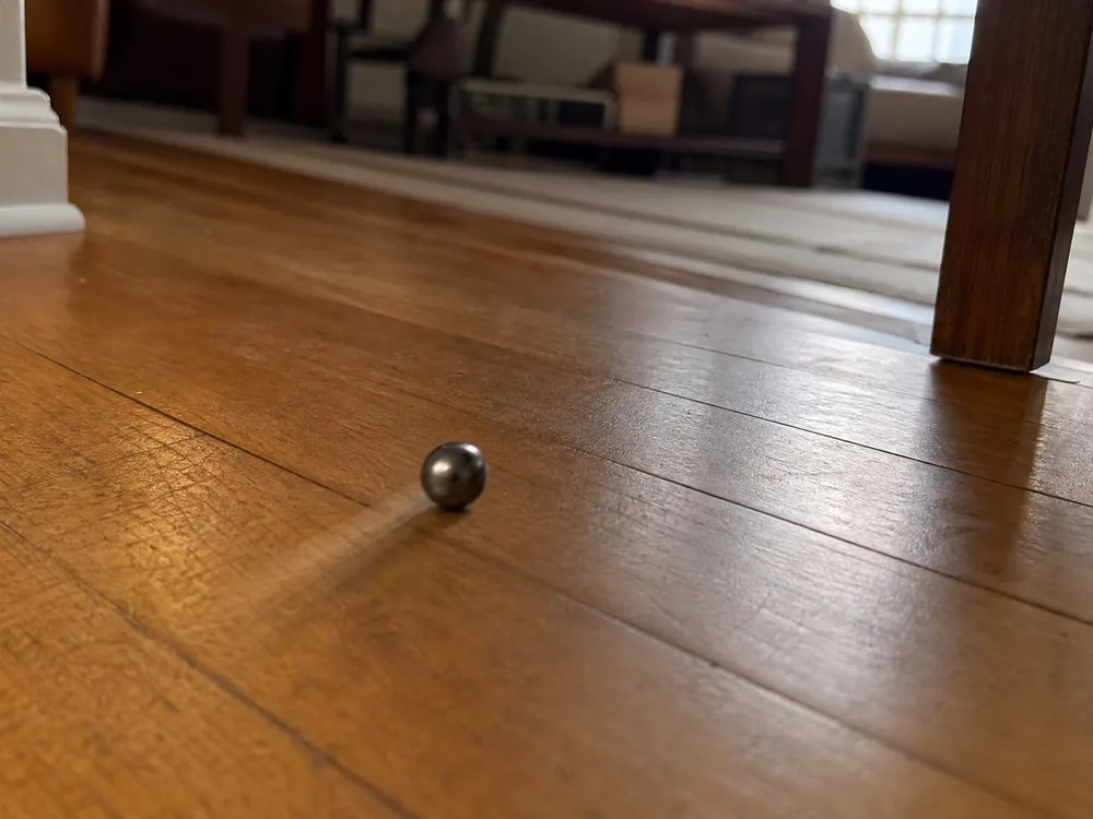 A steel marble rolling across a hardwood living-room floor, blurred by its own travel, showing the floor is off level.